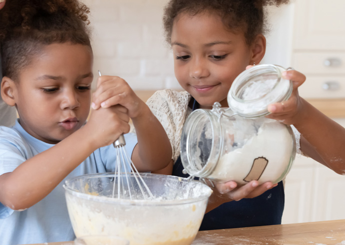 Two children pouring sugar into a bowl of cake mix