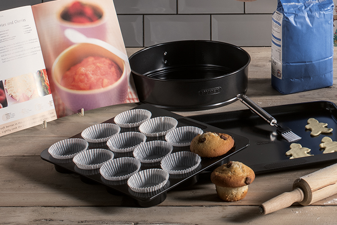Baking trays and cake tins on a wooden countertop with an open recipe book.