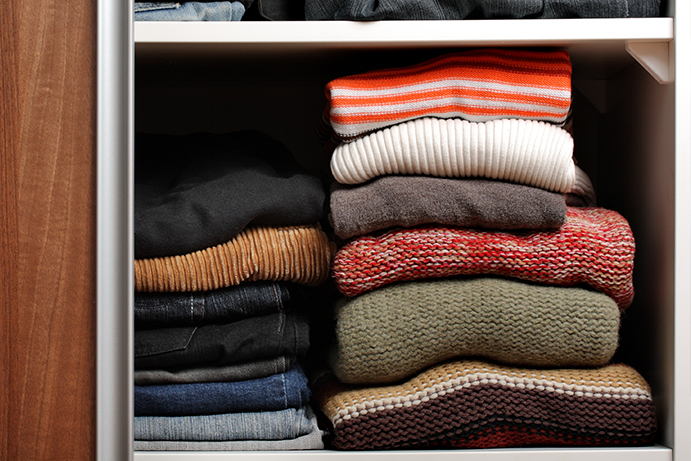Pile of folded jumpers in autumnal colours resting on a white shelf in a dark wood wardrobe.