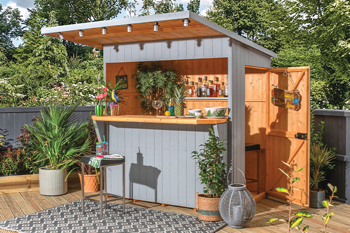 Garden bar shed painted grey, surrounded by plants on outdoor decking in the sunshine