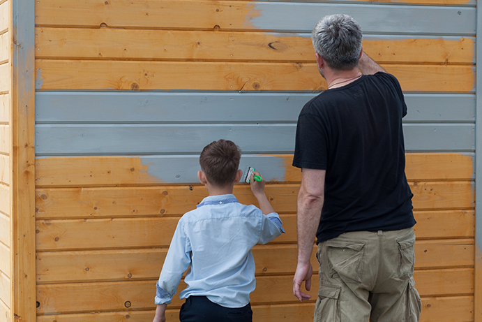 Man and son painting a large wooden buildingwith light grey paint