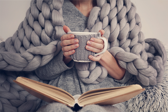 person wrapped in a grey giant knit throw holding a hot cup of tea with a book on their lap