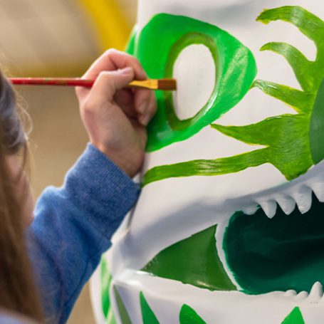 Up close of a hand using a paintbrush to paint onto a white plaster dinosaur sculpture